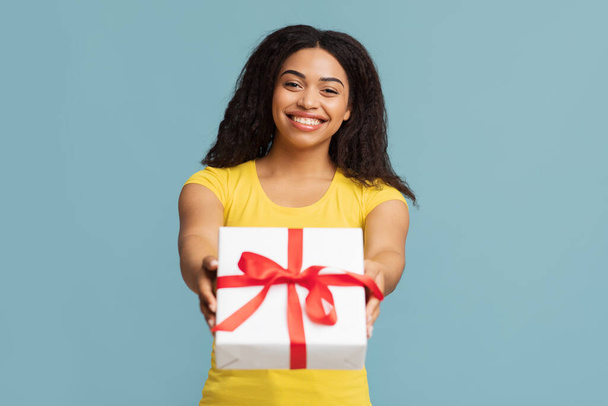Gruß und Feier. Fröhliche schwarze Dame hält verpackten Geschenkkarton in der Hand, streckt Geschenk in die Kamera, blauer Hintergrund - Foto, Bild