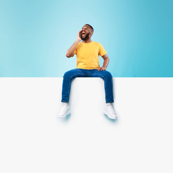 gelukkig zwart man zitten op wit blanco bordje, schreeuwen, kondigen enorme verkoop op blauwe achtergrond, mockup - Foto, afbeelding