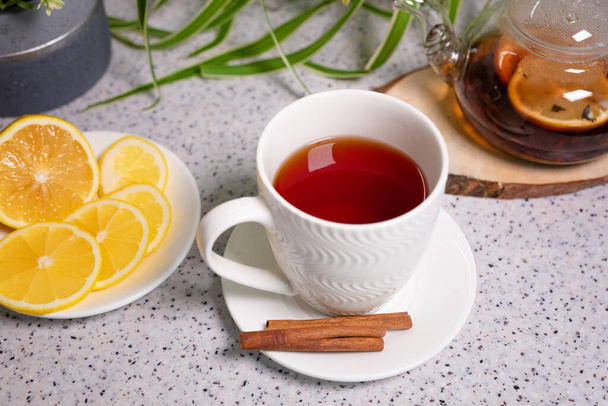 Een warm theedrankje met citroen en sinaasappel, een theepot en een wit kopje, geserveerd met heerlijke citroen en kaneel. Geurige en warme thee - Foto, afbeelding