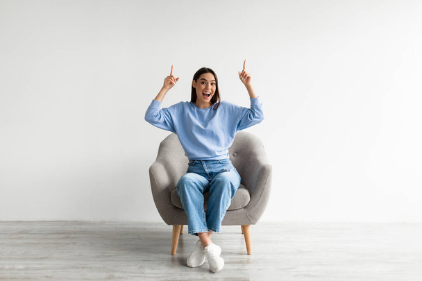 Excited Caucasian woman sitting in armchair, pointing two index fingers up at empty space above her head, mockup - Zdjęcie, obraz