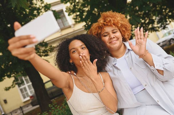 Lovely beautiful happy lesbian African American couple hugging making selfie around city street landscape at summer. LGBT community concept. Female friends smiling enjoying love moments together - Foto, Imagem