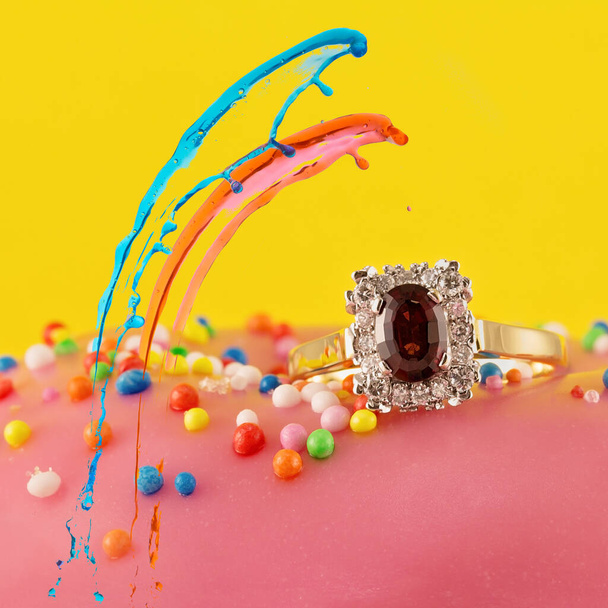 Colored splashes over a garnet solitaire ring surrounded by diamonds in a square shape with gold band on a pink iced cake with sprinkles - Photo, Image