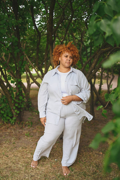 Fashion portrait of young naturally beautiful plus size African American woman in light trouser suit with afro red hair posing in nature parkland in green foliage. Diversity and body positive concept. - Fotó, kép