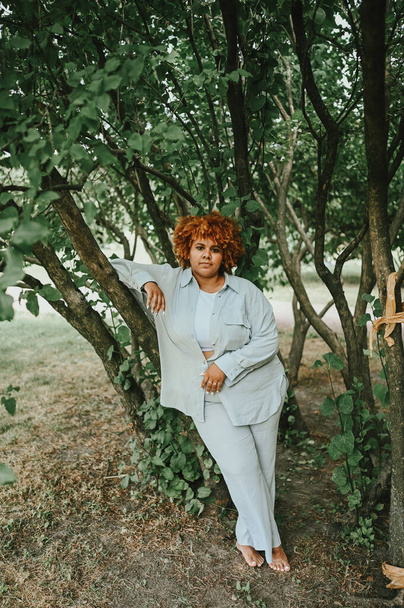 Fashion portrait of young naturally beautiful plus size African American woman in light trouser suit with afro red hair posing in nature parkland in green foliage. Diversity and body positive concept. - Фото, изображение