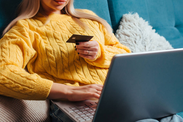 een vrouw met een laptop in een gele trui zit op de bank en rijdt met een bepaalde creditcard. het concept van kopen terwijl je thuis zit. - Foto, afbeelding