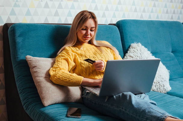 una donna con un computer portatile in un maglione giallo siede sul divano e guida una determinata carta di credito. il concetto di acquisto mentre si è seduti a casa. - Foto, immagini