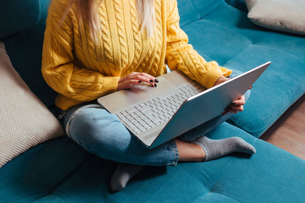 a woman with a laptop in a yellow sweater is sitting on the couch . the concept of remote work and shopping while sitting at home. - Zdjęcie, obraz