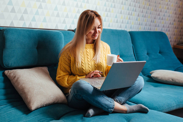Egy nő egy laptoppal sárga pulóverben ül a kanapén. a távmunka és a távvásárlás fogalma otthon ülve. - Fotó, kép