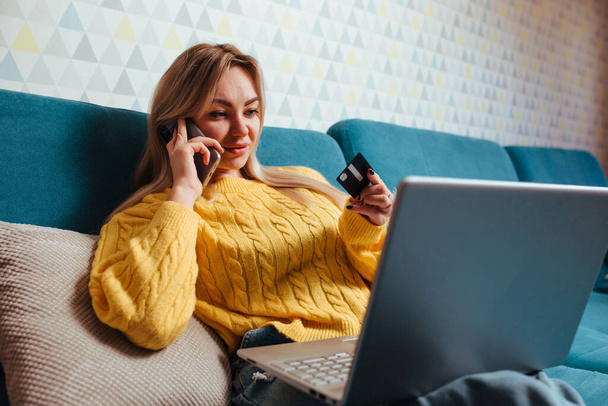 Sarı kazaklı dizüstü bilgisayarı olan bir kadın kanepeye oturur ve kredi kartı kullanır. Evde otururken satın alma kavramı. - Fotoğraf, Görsel