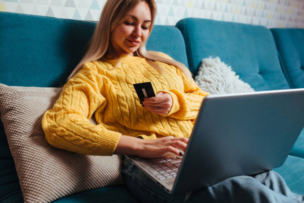 een vrouw met een laptop in een gele trui zit op de bank en rijdt met een bepaalde creditcard. het concept van kopen terwijl je thuis zit. - Foto, afbeelding