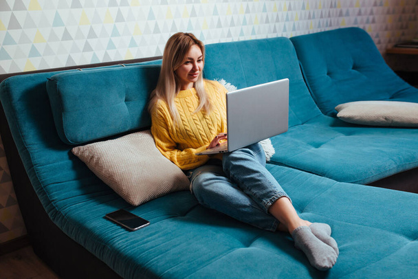 une femme avec un ordinateur portable dans un pull jaune est assise sur le canapé. le concept de télétravail et de magasinage assis à la maison. - Photo, image