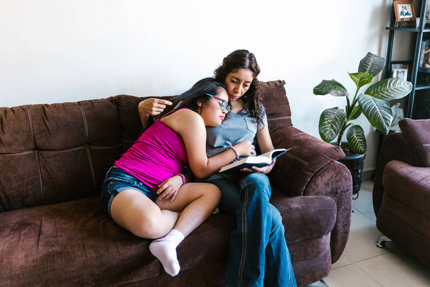Latin mother reading a book to her teenage daughter with down syndrome sitting on the sofa at home, in disability concept in Latin America - Photo, Image