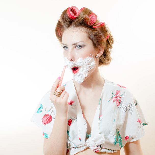 Beautiful young pinup woman shaving face with foam and razor looking in camera isolated on white background closeup portrait - Fotó, kép
