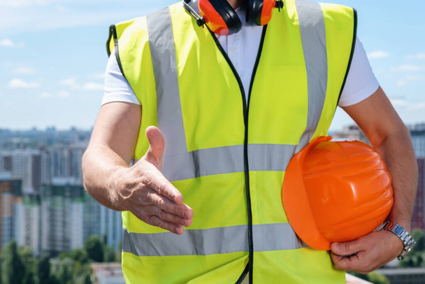 Builder holds out one hand for a handshake and the other holds an orange helmet - Photo, Image