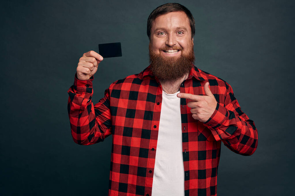 Портрет веселого бородатого мужчины, указывающего пальцем на кредитку на сером фоне. - Фото, изображение