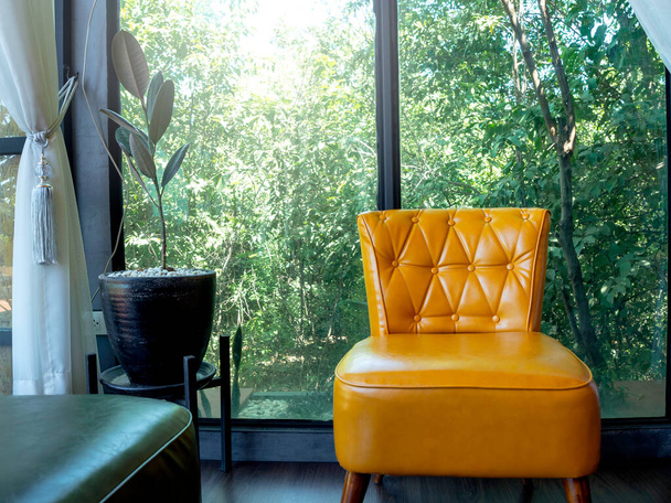 Gelbe Vintage-Ledercouchbank mit Anstecknadeln und Knöpfen Dekoration in der Nähe von Pflanzentopf und Glasfenster mit Blick auf die grüne Natur draußen. - Foto, Bild