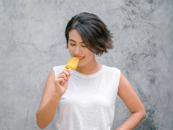 Frauen essen Eis am Stiel. Glückliche Asiatin mit kurzen Haaren im lässigen weißen ärmellosen Hemd, das im Sommer gelbes Eis am Stiel auf grauem Betonwandhintergrund isst. - Foto, Bild