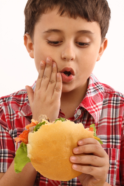 Poika syö ison hampurilaisen läheltä.
 - Valokuva, kuva