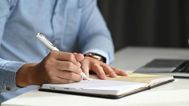 Zavřít pohled na obchodníka držícího pero při psaní do sešitu a sezení na pracovišti. - Fotografie, Obrázek