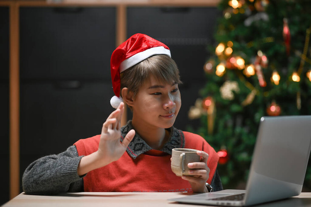 Lächelnde fröhliche Frau mit Weihnachtsmütze, Kaffeetasse und Videotelefonie mit ihren Freunden und winkender Hand zu Hause. - Foto, Bild