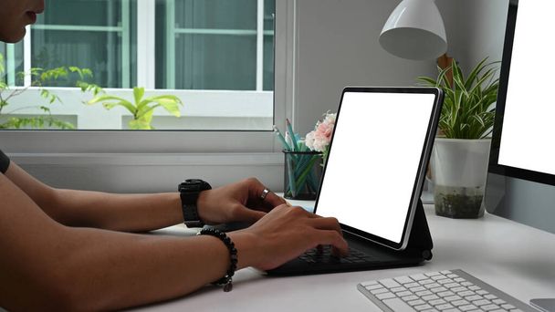 Crop shot του συμπυκνωμένο νεαρός ελεύθερος επαγγελματίας δακτυλογραφεί σε tablet υπολογιστή με λευκή οθόνη, ενώ κάθεται στο χώρο εργασίας. - Φωτογραφία, εικόνα
