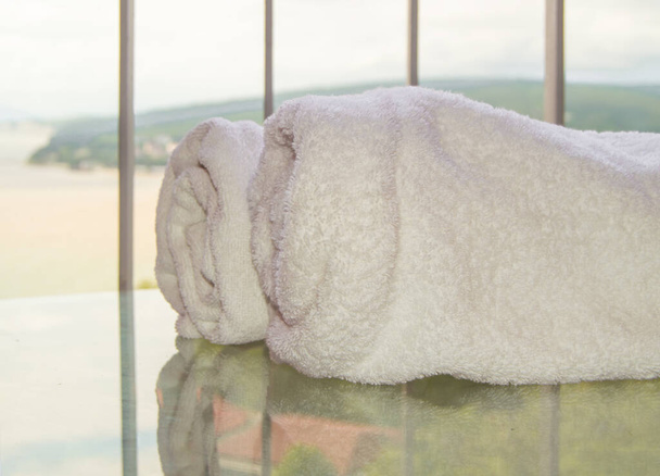 Zbliżenie dwóch rolek białych ręczników frotte leżących na przezroczystym szklanym stole na balkonie, efekt odbicia. - Zdjęcie, obraz