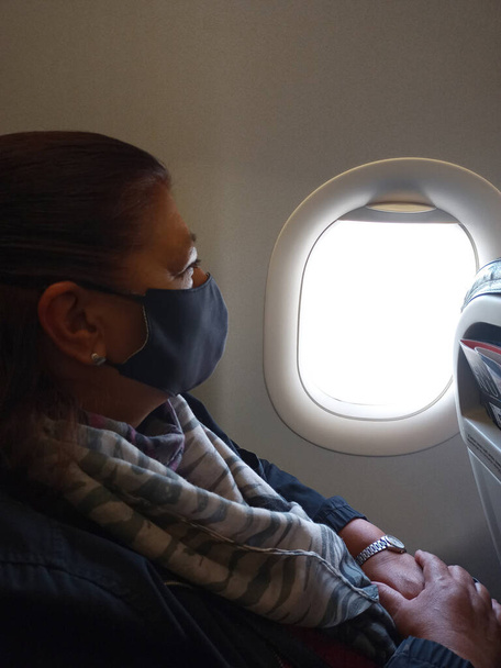 reife Latina im Flugzeug, die wegen der Pandemie mit Gesichtsmaske aus dem Fenster schaut - Foto, Bild