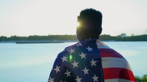 Uomo afro-americano con bandiera americana sulle spalle guarda in lontananza all'alba - Filmati, video