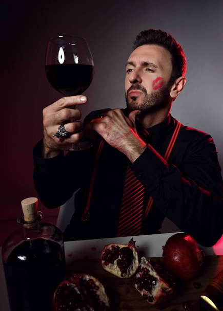 Brutal man in black shirt and trace of kiss on cheek sits at table with pomegranate and juice and loks at glass in hand - Photo, image