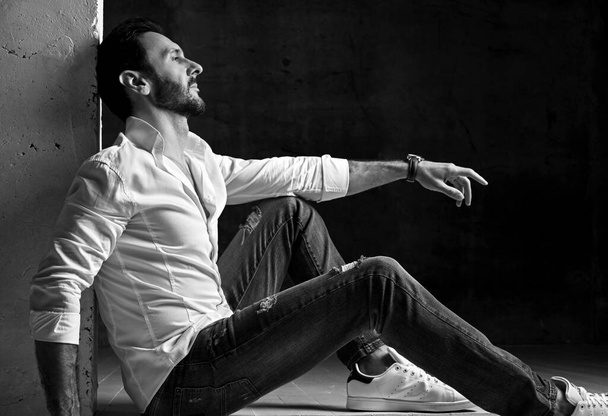 Black and white. Tired unshaved man in white shirt and jeans sits leaning on concrete wall is resting and relaxing - Photo, Image