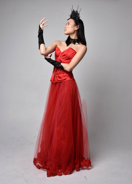 Full length  portrait of beautiful young asian woman wearing red corset and ornate gothic queen crown. Graceful standing posing  isolated on studio background. - Photo, Image
