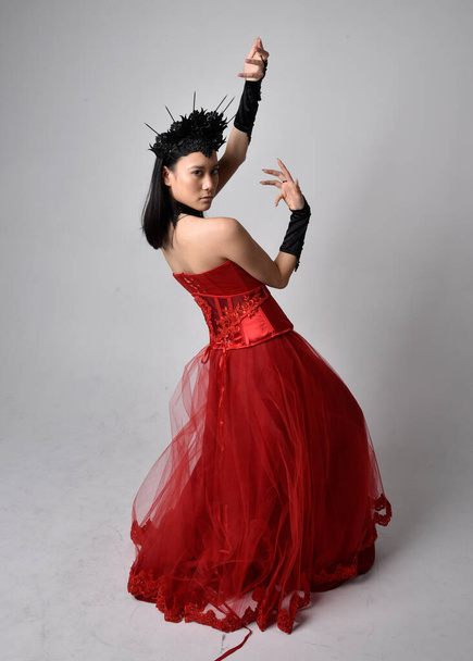 Full length  portrait of beautiful young asian woman wearing red corset and ornate gothic queen crown. Graceful standing posing  isolated on studio background. - 写真・画像