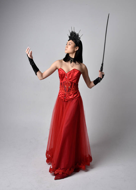 Full length  portrait of beautiful young asian woman wearing red corset and ornate gothic queen crown. Graceful standing posing  isolated on studio background. - Фото, изображение