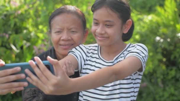 Iloinen suloinen teini lapsenlapsi ja hänen vanhempi isoäitinsä ottavat selfie-valokuvan matkapuhelimella ja hymyilevät katsoessaan kameraa kotona puutarhassa. Suhde perheeseen. - Materiaali, video