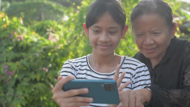 Χαρούμενη αξιολάτρευτη έφηβη κοπέλα και η ηλικιωμένη γιαγιά της απολαμβάνουν με συνομιλία μέσω βίντεο στην οικογένειά τους από κινητό smartphone στον κήπο στο σπίτι. Σχέση δεσμού στην οικογένεια. - Πλάνα, βίντεο