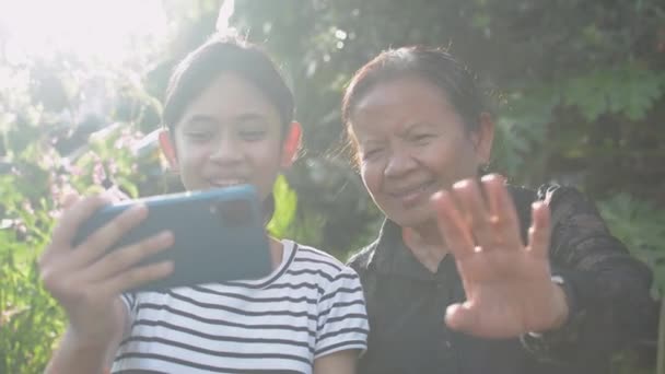 Asyalı sevimli genç kız ve büyük annesi evlerinin arka bahçesindeki cep telefonundan aileleriyle görüntülü sohbet ederken el sallamaktan zevk alıyorlar. Aile bağları. - Video, Çekim