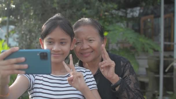 Mutlu Asyalı sevimli genç kız ve kıdemli büyükannesi sabah güneşinin altında cep telefonlarıyla evlerinin önünde selfie çekiyorlar. Ailedeki iki nesli birbirine bağlamak. - Video, Çekim