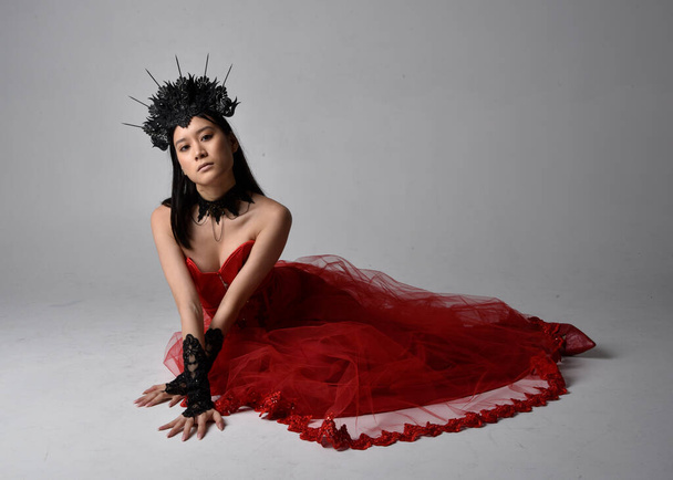 Portrait pleine longueur de belle jeune femme asiatique portant corset rouge et couronne de reine gothique ornée. Assis gracieux posant isolé sur fond de studio. - Photo, image
