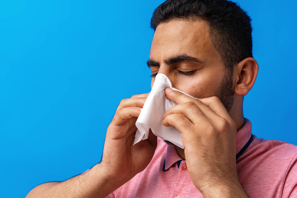 Young man in pink shirt with allergy or cold, blowing his nose in a tissue against blue background - 写真・画像