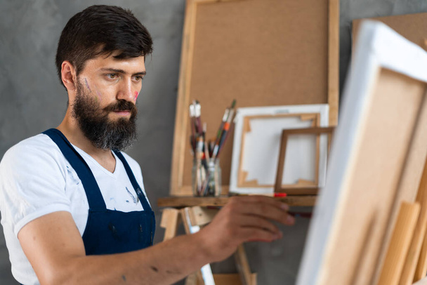 Nahaufnahme eines bärtigen männlichen Künstlers in einer schmutzigen Schürze steht in einem Kunstatelier, hält einen Pinsel in der Hand und malt ein Bild. - Foto, Bild