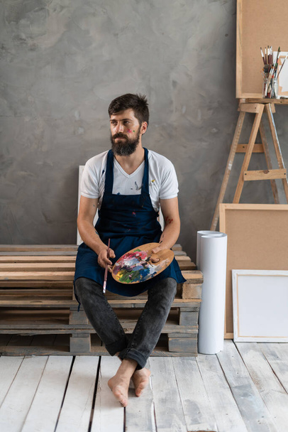 Ένας γενειοφόρος άντρας καλλιτέχνης ντυμένος με μπλε βρώμικη ποδιά κάθεται σε παλέτες σε ένα στούντιο τέχνης και ξεκουράζεται. - Φωτογραφία, εικόνα