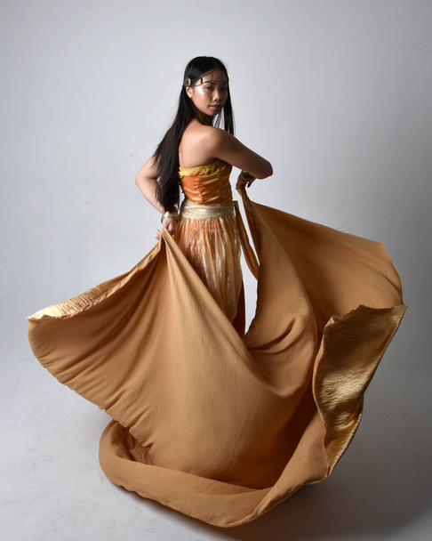 Retrato de larga duración de una hermosa joven asiática vestida con túnicas árabes doradas como un genio, de pie posando sosteniendo tela fluida, aislada en el fondo del estudio. - Foto, Imagen