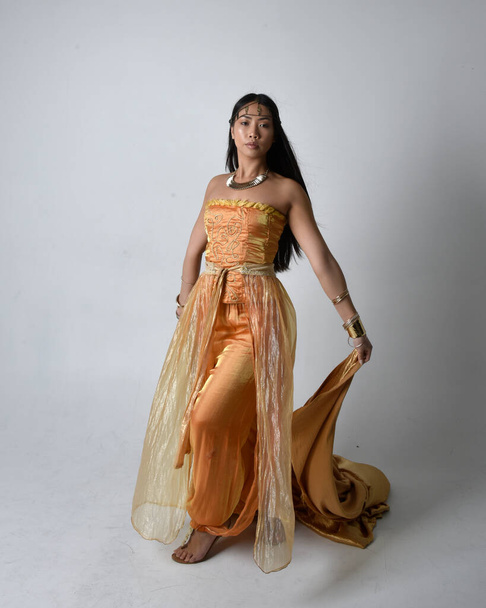 Retrato de comprimento total de mulher asiática muito jovem vestindo vestes árabes douradas como um gênio, em pé pose segurando tecido fluindo, isolado no fundo do estúdio. - Foto, Imagem