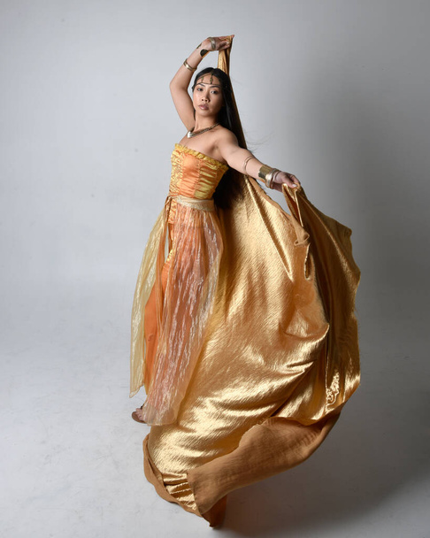 Altın Arap cübbesi giymiş güzel Asyalı bir kadının tam boy portresi. Stüdyo arka planında izole edilmiş, akan kumaşı tutan bir cin gibi duruyor.. - Fotoğraf, Görsel