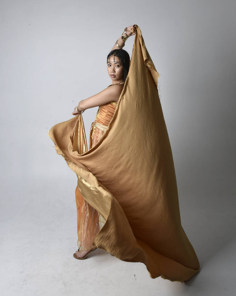 Full length portrait of pretty young asian woman wearing golden Arabian robes like a genie, standing pose holding flowing fabric, isolated on studio background. - Foto, afbeelding