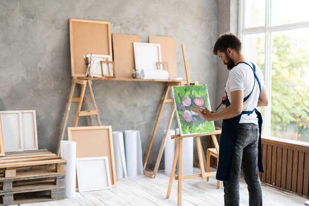 σε ένα δημιουργικό στούντιο τέχνης, ένας γενειοφόρος καλλιτέχνης στέκεται κρατώντας ένα πινέλο και μια παλέτα με χρώματα, ζωγραφίζει μια ελαιογραφία σε καμβά με ανθισμένες παιώνιες. Αντιγραφή χώρου. - Φωτογραφία, εικόνα