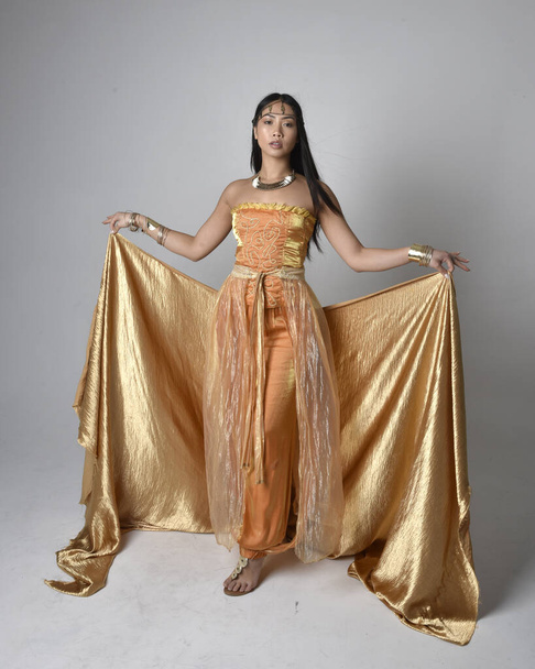 Portret pięknej młodej azjatyckiej kobiety w złotych arabskich szatach, jak dżin, stojącej w pozycji stojącej z płynącą tkaniną, odizolowanej na tle studia. - Zdjęcie, obraz