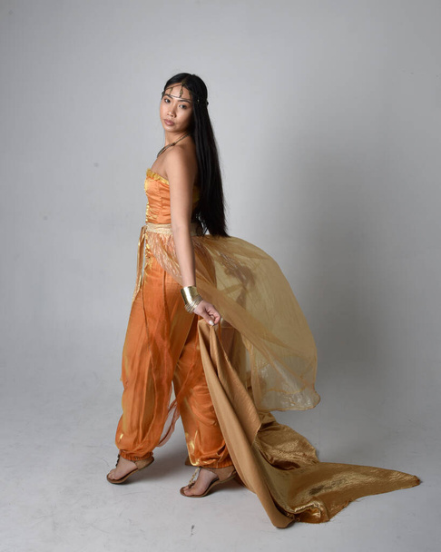 Retrato de larga duración de una hermosa joven asiática vestida con túnicas árabes doradas como un genio, de pie posando sosteniendo tela fluida, aislada en el fondo del estudio. - Foto, Imagen
