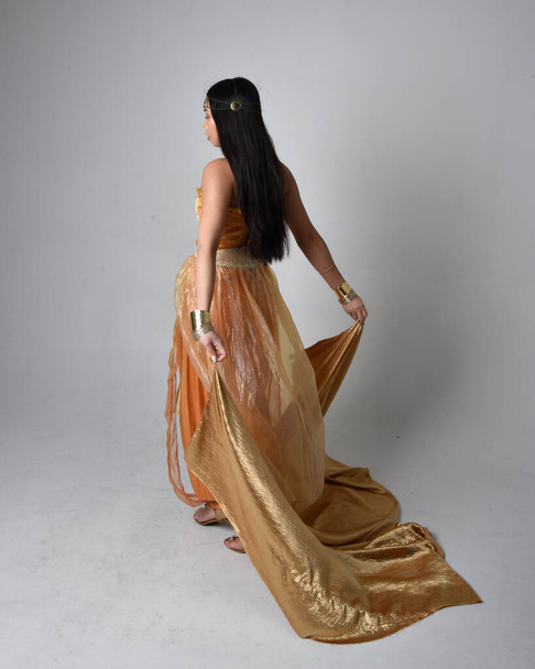 ジーニーのような黄金のアラビア語のローブを身に着けているかなり若いアジアの女性の完全な長さの肖像画,流れるような布を保持立ってポーズ,スタジオの背景に孤立. - 写真・画像