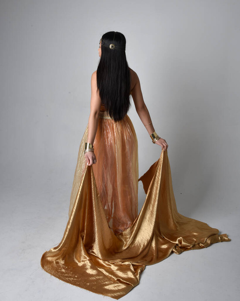 Teljes hossz portré csinos fiatal ázsiai nő visel arany arab köntös, mint egy dzsinn, álló póz gazdaság áramló szövet, elszigetelt stúdió háttér. - Fotó, kép